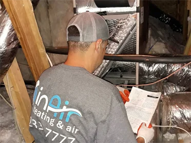 A technician wearing a company shirt examines home improvement HVAC system components in an attic, surrounded by insulation and wooden beams.