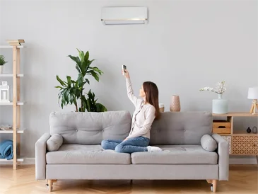 A woman sitting on a gray sofa in a home decor-inspired living room, taking a selfie with her smartphone.