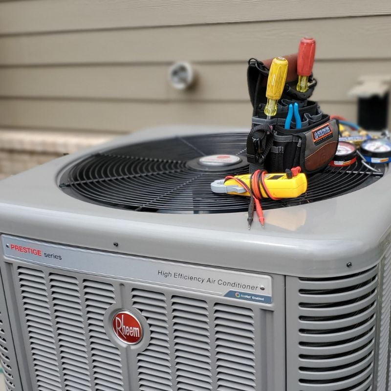 A tool bag and various HVAC repair tools sit atop a high-efficiency Rheem air conditioning unit outside a house.