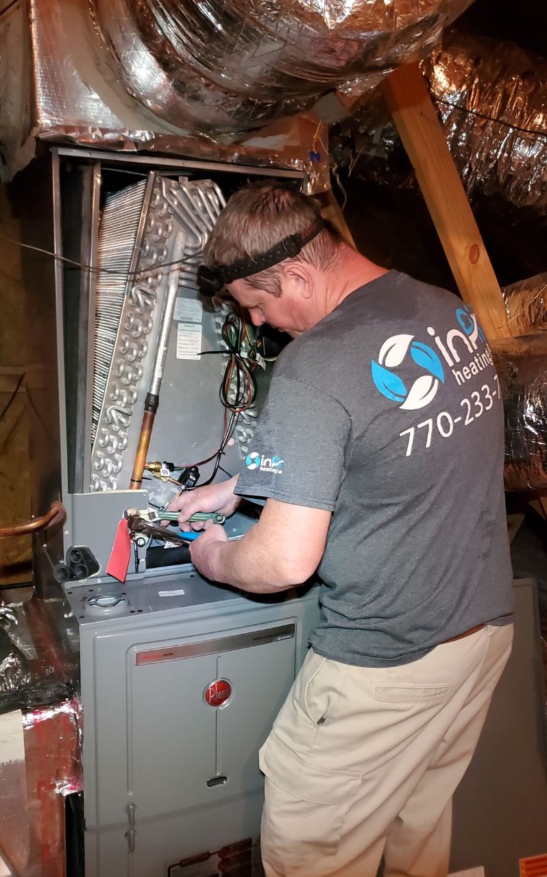 A technician in a branded shirt services an HVAC unit in a cramped attic space, using tools on the electronic components.