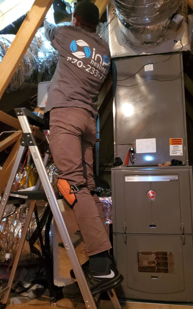 A technician in a branded shirt services HVAC ducts in an attic, standing on a ladder close to installed equipment.