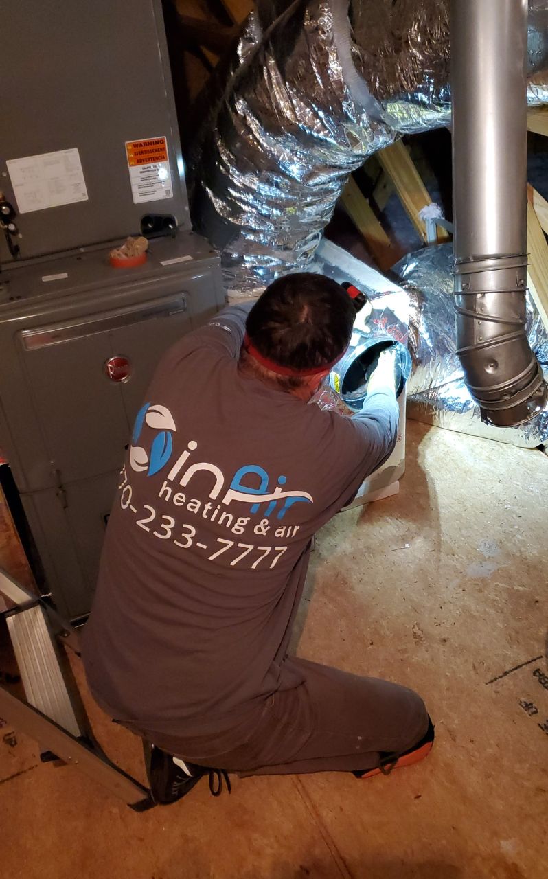 Man in a branded uniform using a tool to work on an HVAC system in a basement area with ductwork visible.