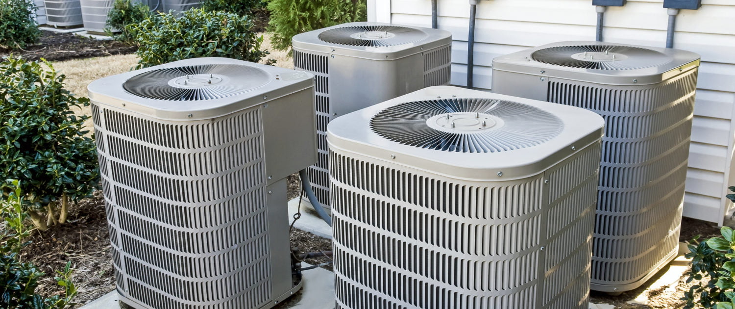 Four residential central air conditioning units outside a home, surrounded by bushes and a white siding wall.