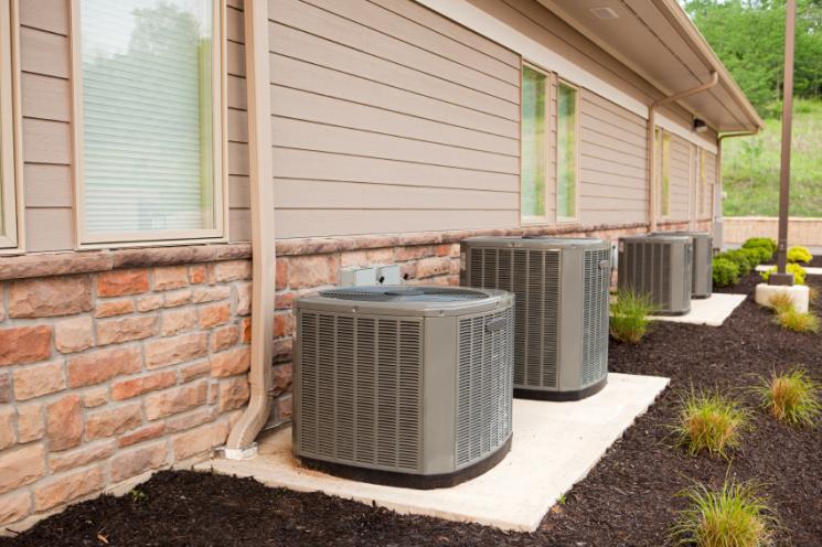 Three air conditioning units beside a residential house with beige siding and stone accents, surrounded by a landscaped garden.