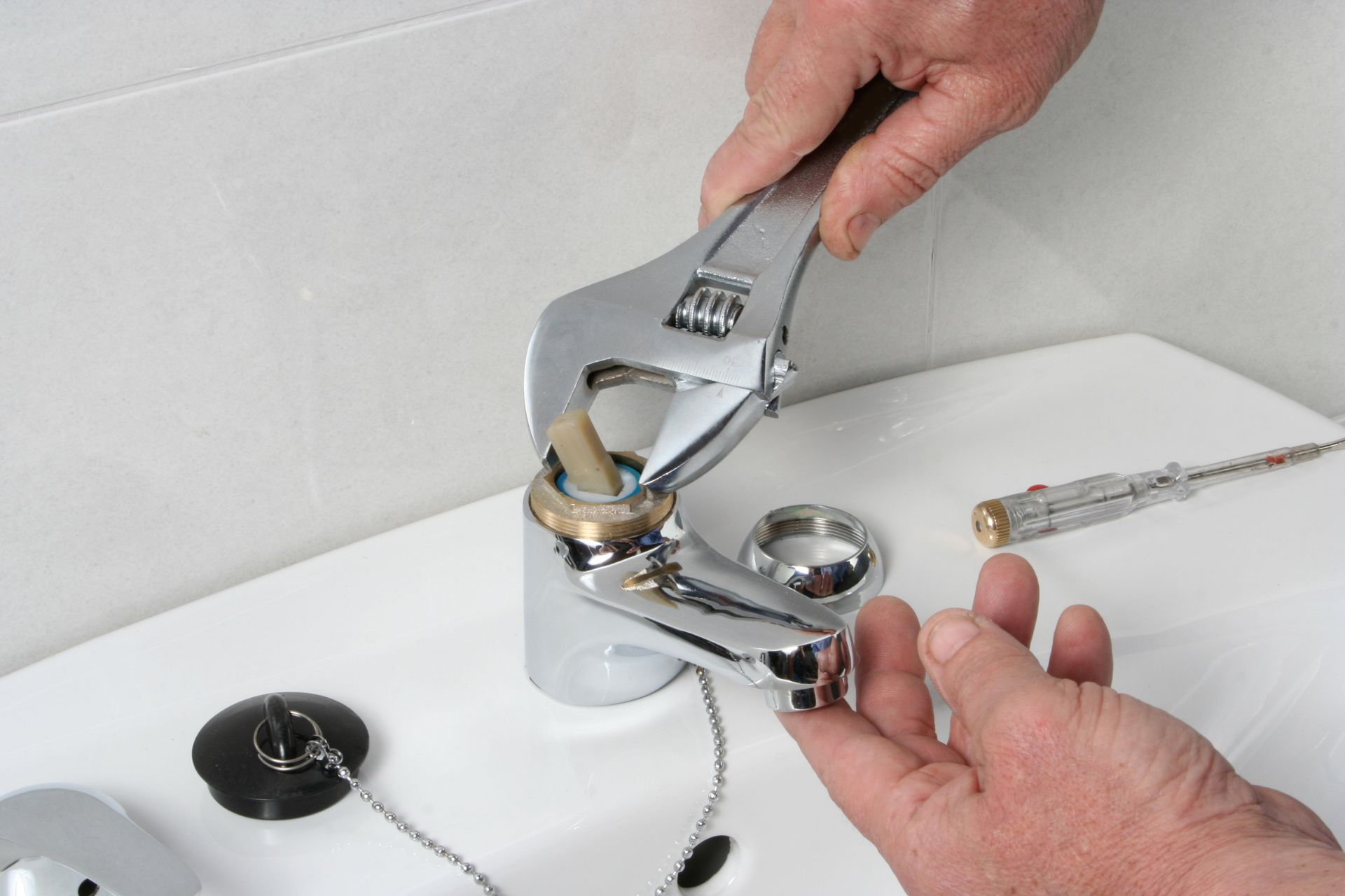 A person uses an adjustable wrench to repair a bathroom faucet, showcasing their plumbing services expertise, with a separate part held in one hand and a screwdriver lying nearby.