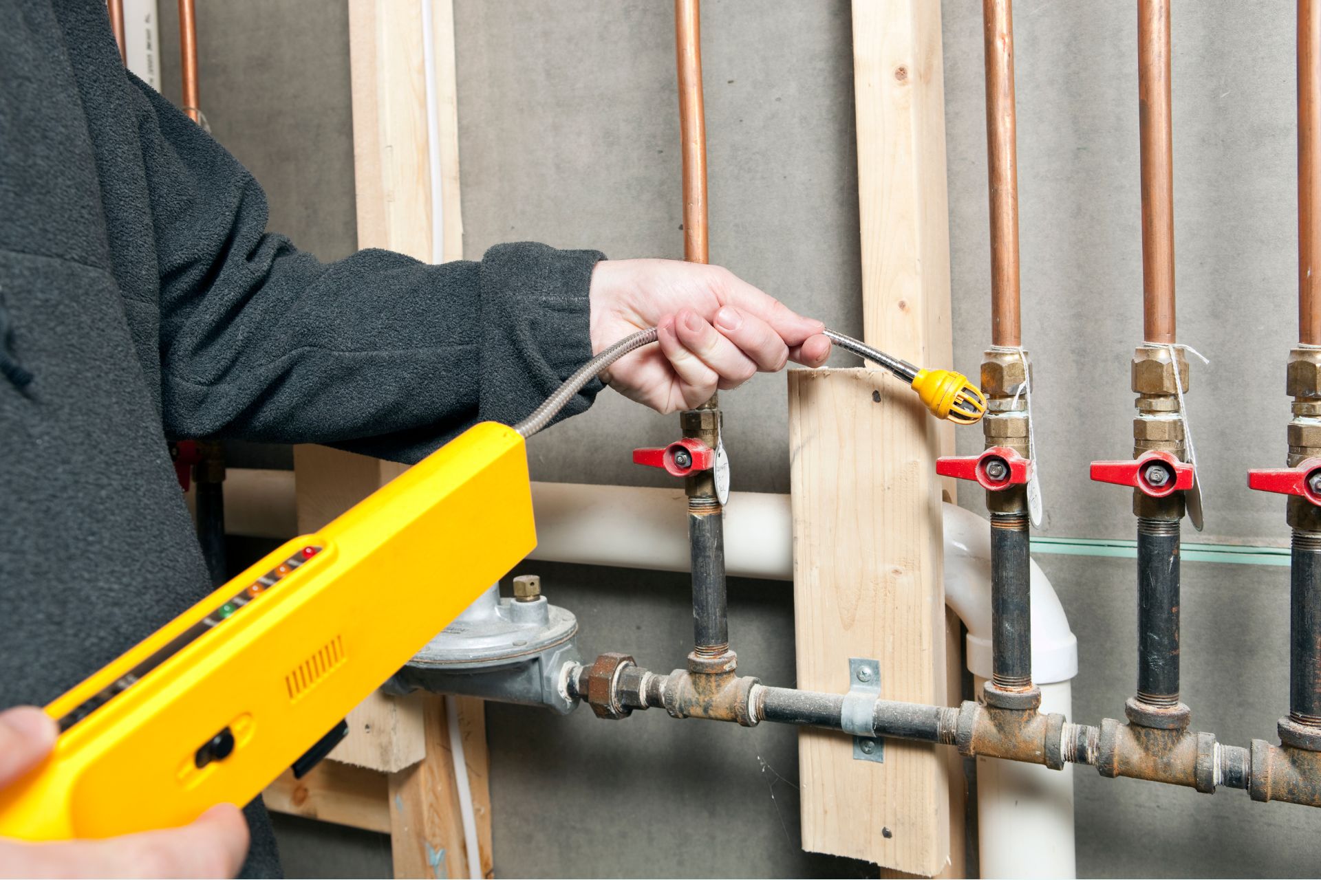 A person using a yellow electronic device to check copper pipes in an unfinished wall with multiple valves, demonstrating precise plumbing services.