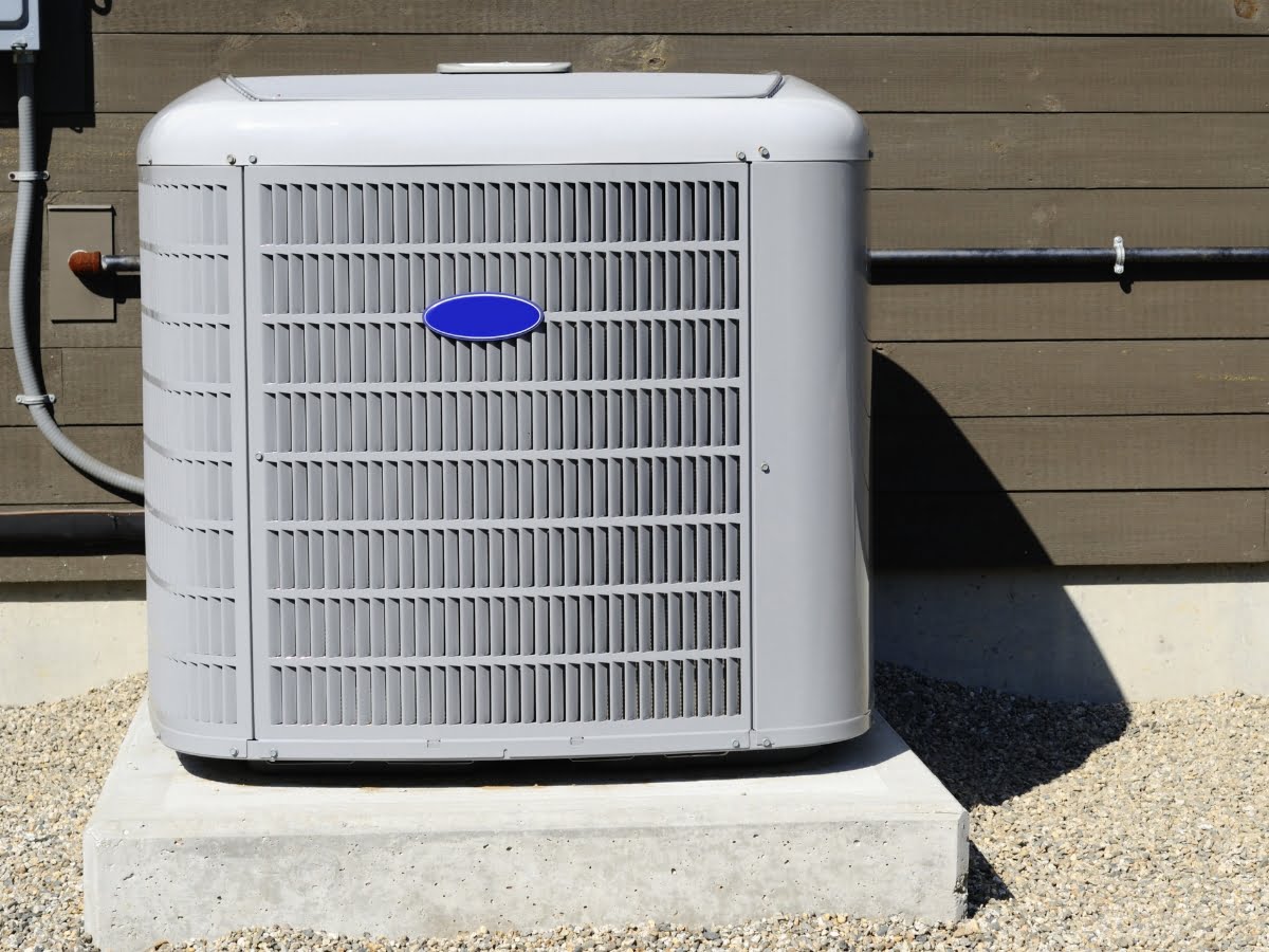 A gray outdoor air conditioning unit, designed to prevent issues like why do heat pumps freeze up, is mounted on a concrete pad against a brown wooden wall.