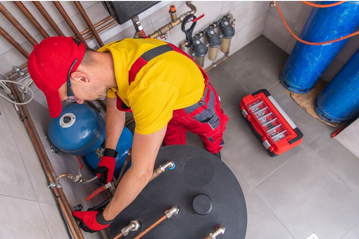 A technician in a red cap and uniform works on a boiler system, adjusting pipes. Nearby is a red toolbox and blue tanks, indicating a maintenance setting. He meticulously installs hot water recirculation systems to ensure efficient water flow throughout the building.