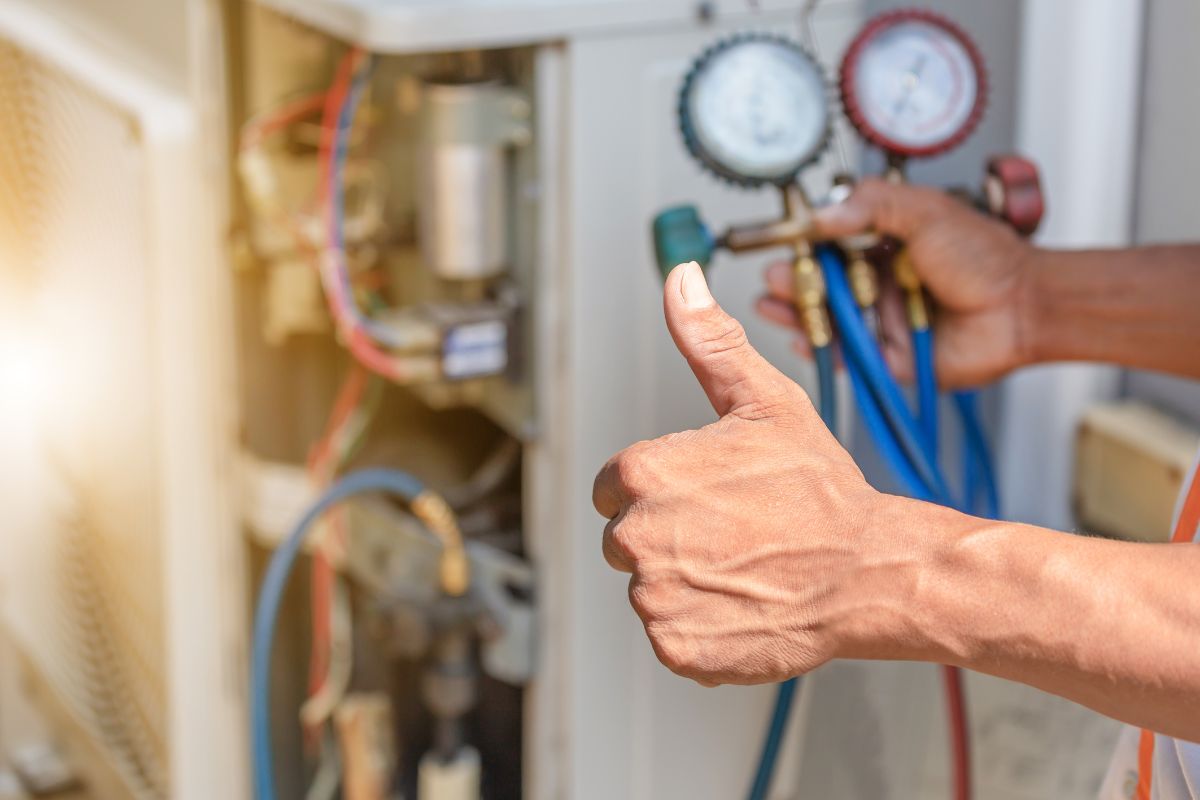 A person gives a thumbs-up while adjusting gauges on an air conditioning unit with hoses attached, ensuring everything is in top shape for the furnace maintenance checklist.