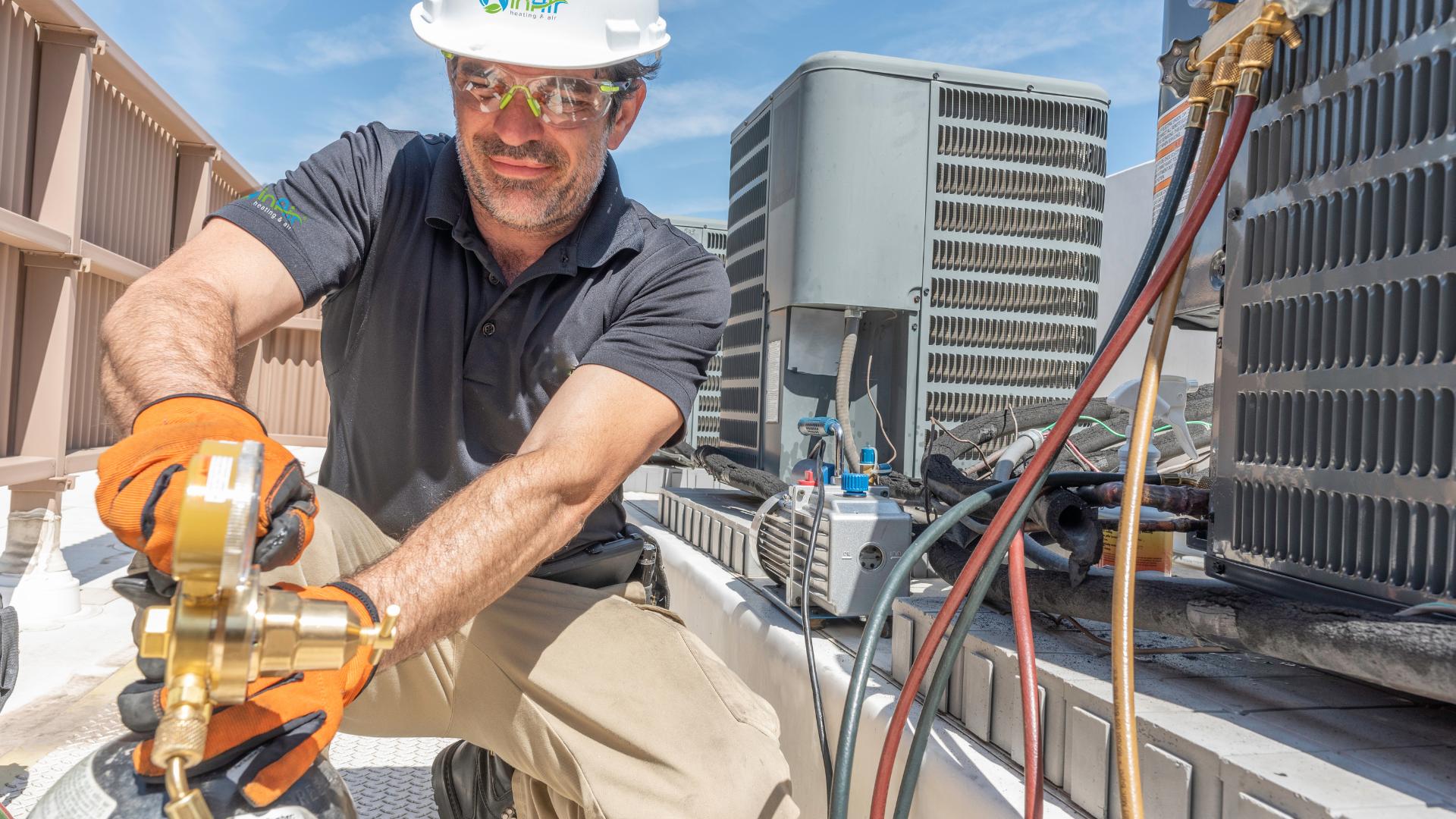 A technician wearing safety gear adjusts a valve on a rooftop HVAC system under a blue sky.