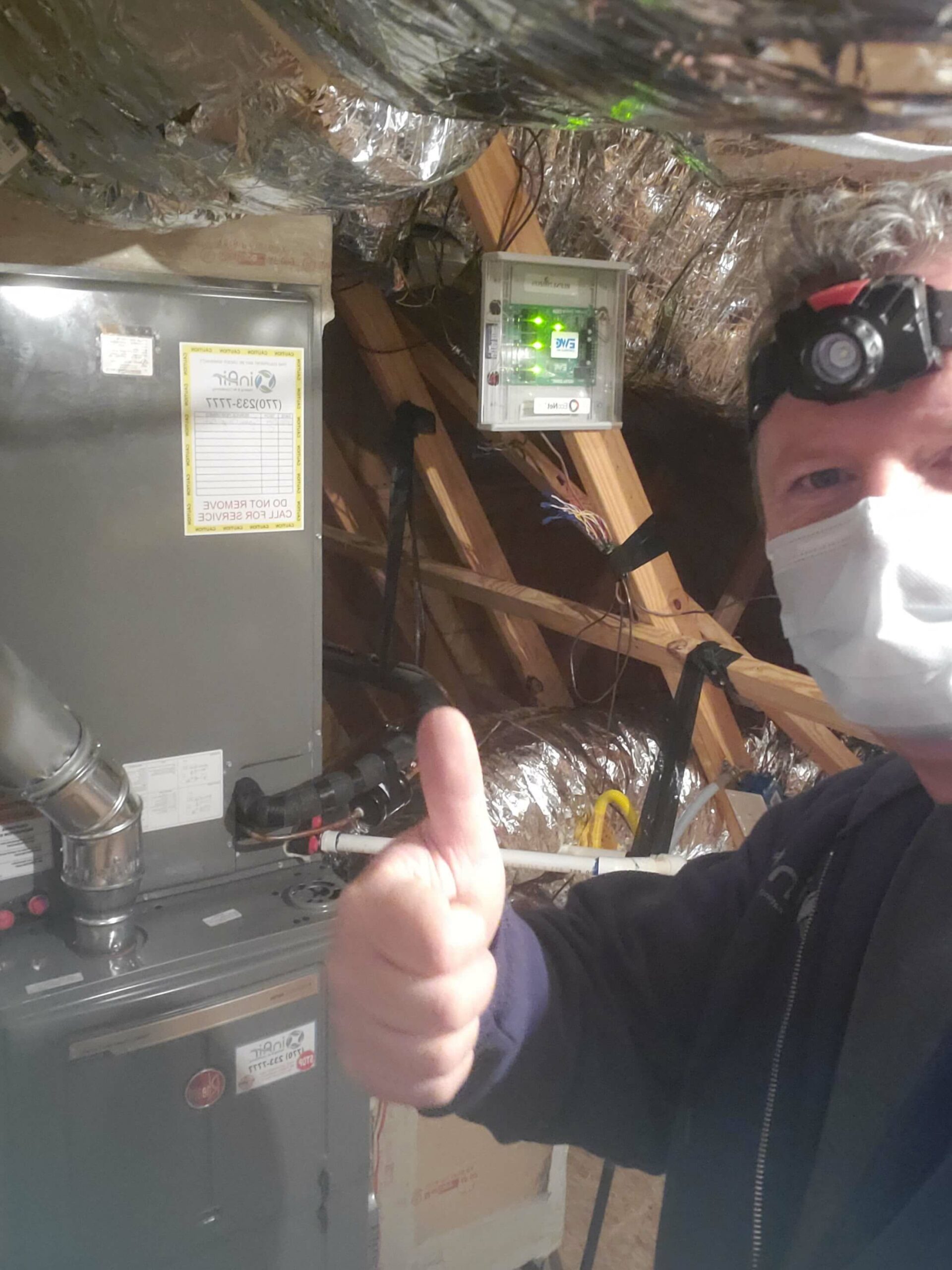 A person wearing a headlamp and face mask gives a thumbs-up gesture in front of an HVAC system inside an attic.