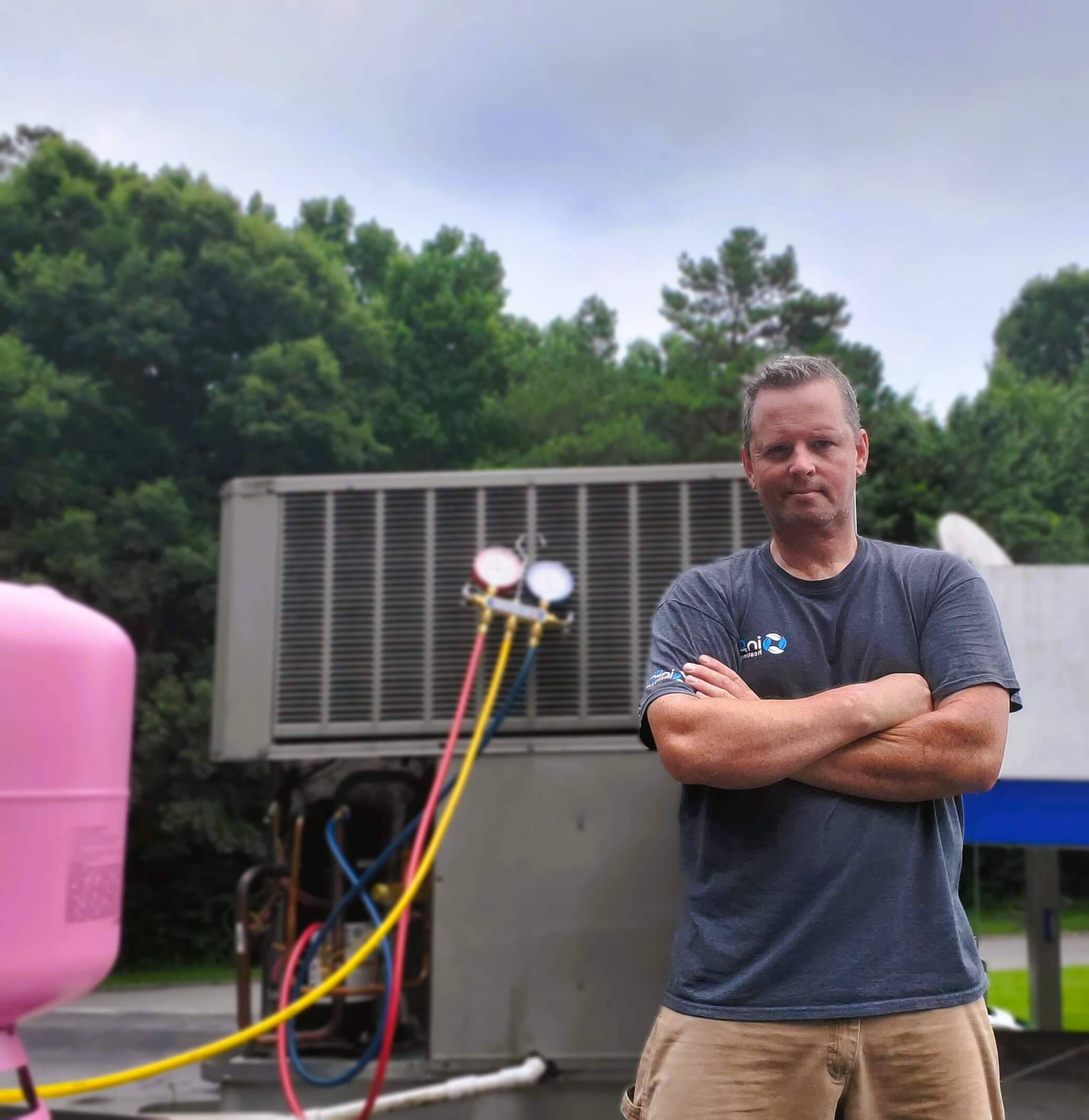 An hvac technician stands with arms crossed in front of an outdoor air conditioning unit. Various gauges and colored hoses are visible beside him used to provide hvac services and preventative maintenance plans.