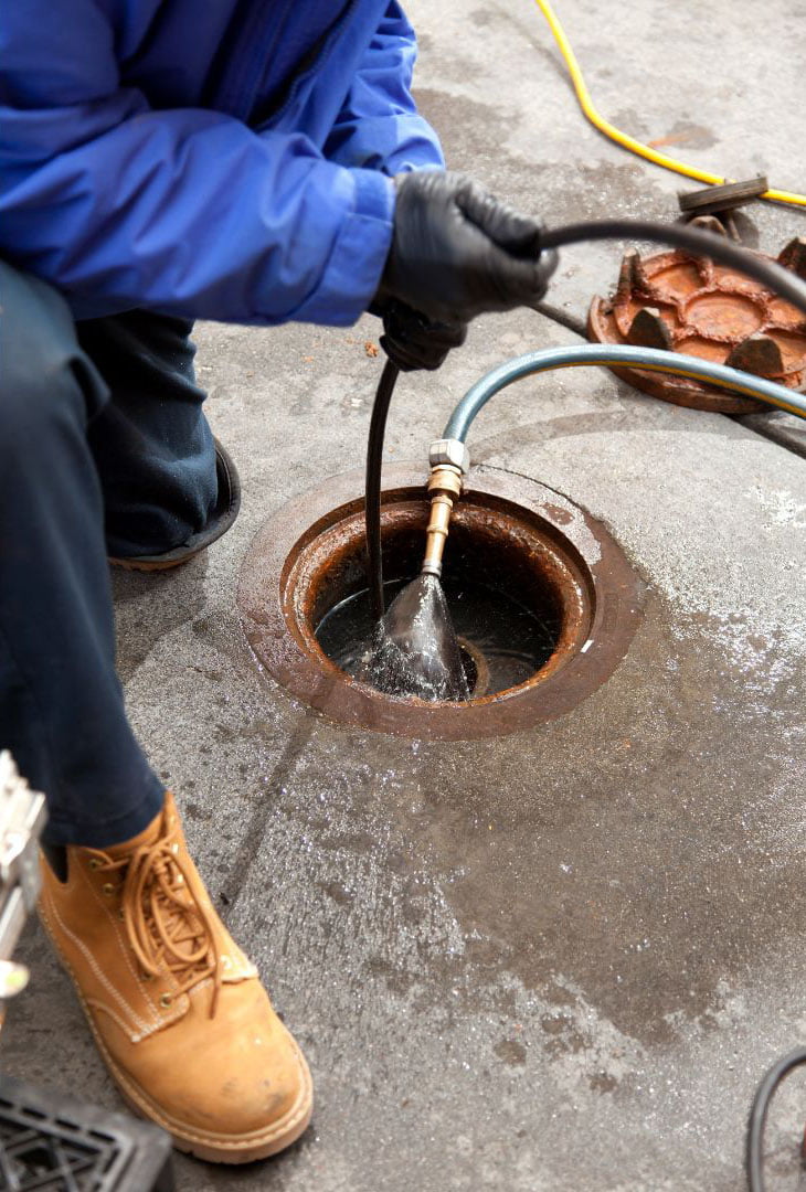 A plumber wearing blue coveralls and gloves uses a high-pressure water jet to clean a sewer manhole.