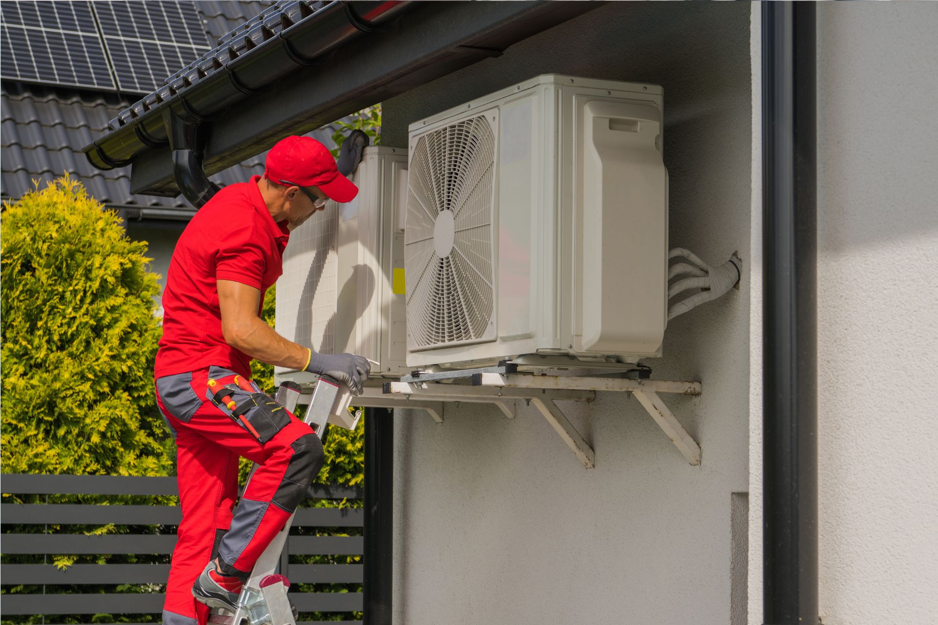 Technician in red uniform standing on ladder, performing heat pump repair on an outdoor air conditioning unit mounted on the exterior wall of a house.