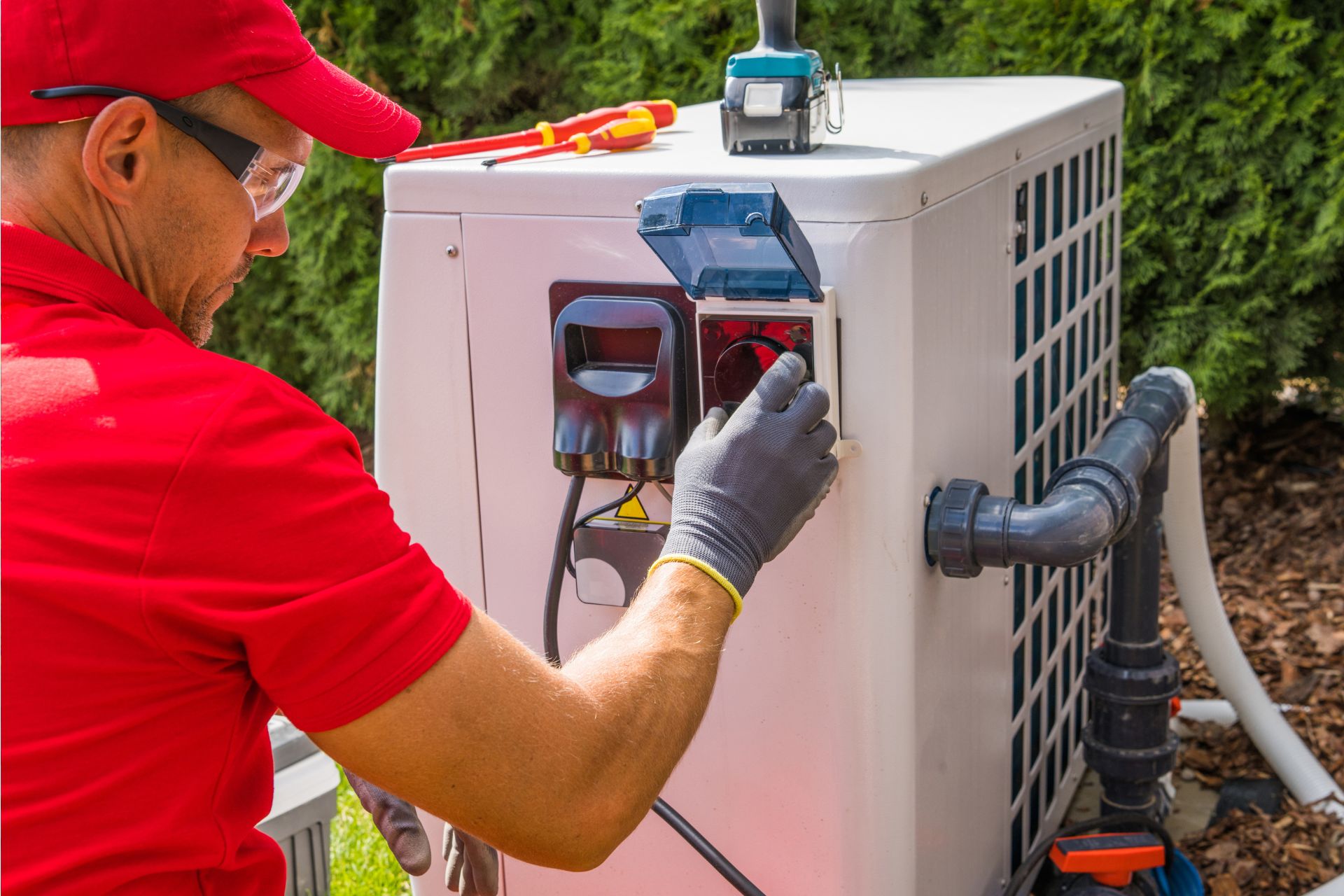 Technician servicing an outdoor air conditioning unit, adjusting a component with tools on top of the unit. The technician, specializing in heat pump repair, is wearing a red shirt, cap, and protective gloves.