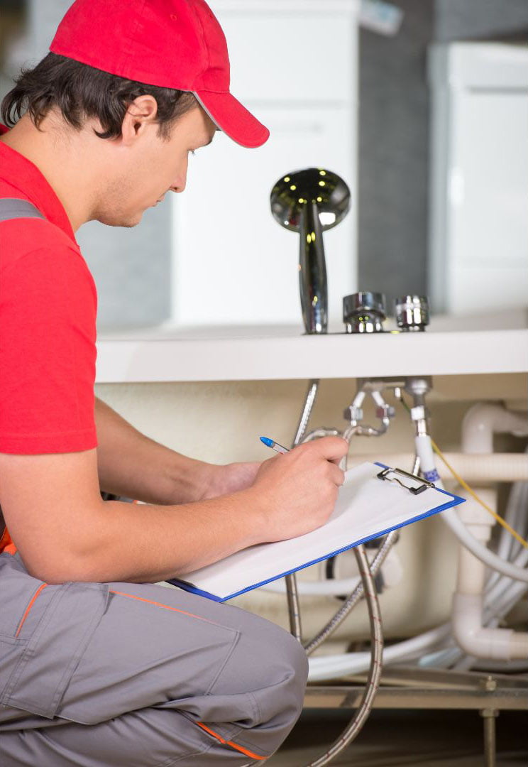 A plumber in a red shirt and cap sitting and writing on a clipboard, with sink pipes visible in the background, ready to provide plumbing services.