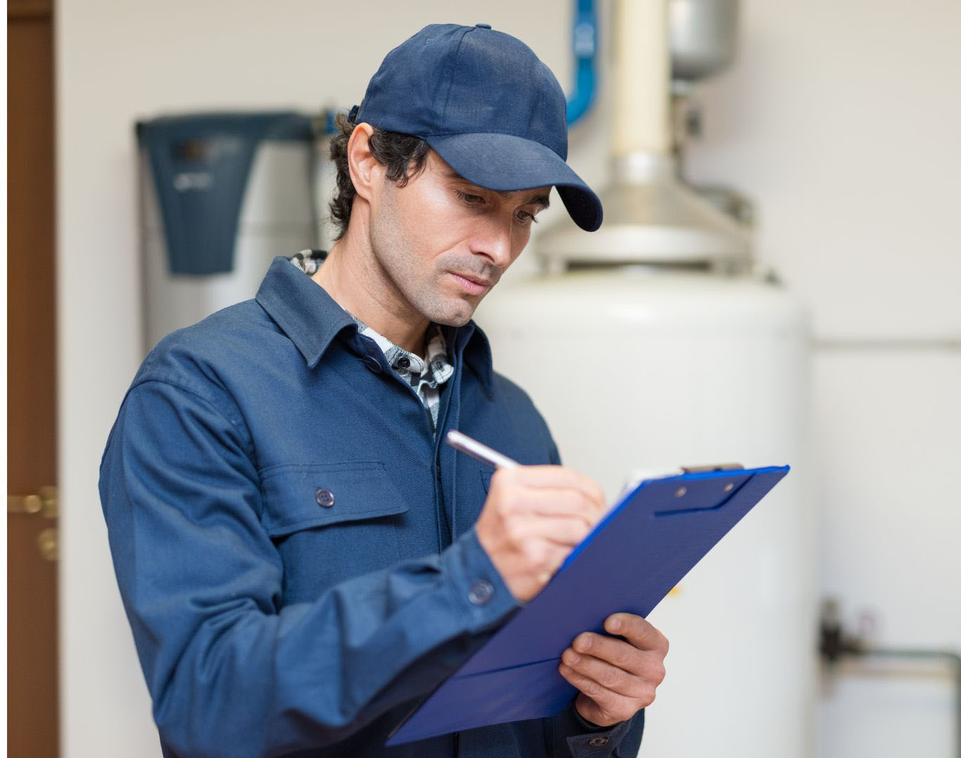 A plumber in a blue uniform and cap taking notes on a clipboard, standing beside a large water heater.