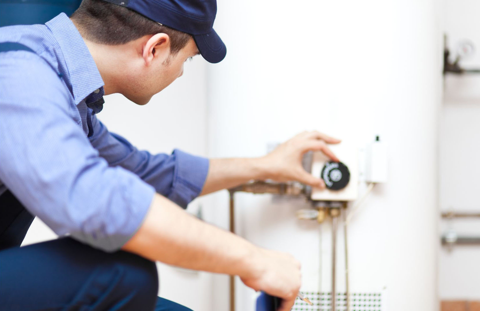 A plumber in a blue uniform performing plumbing repair on the thermostat of a water heater.