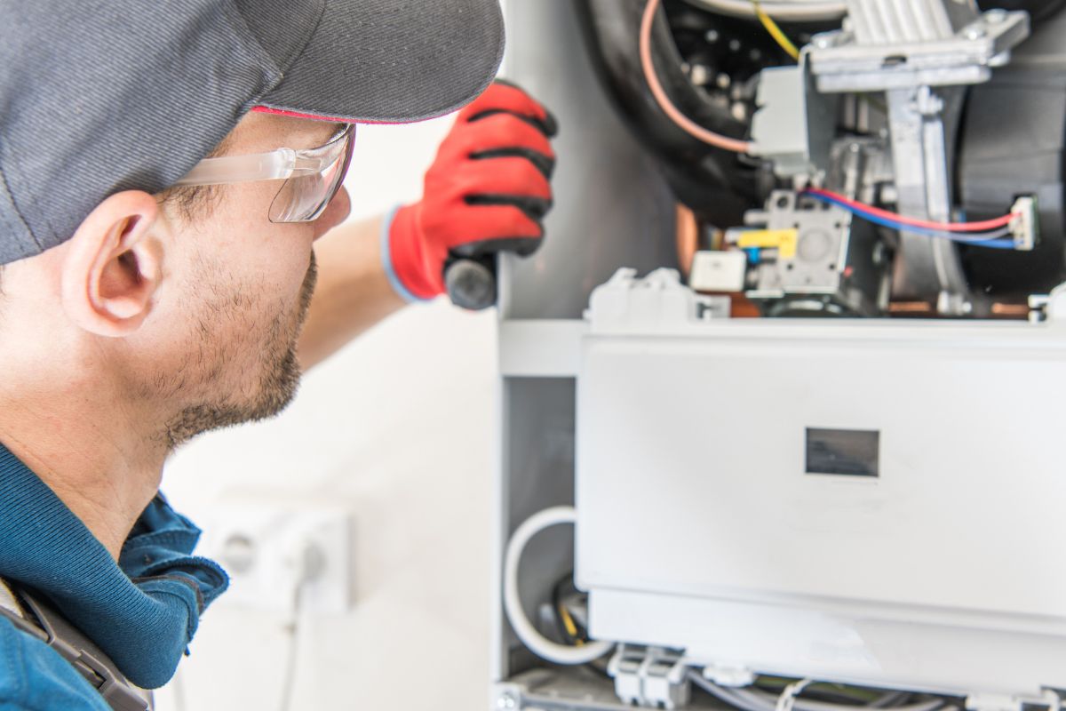 A technician in safety glasses and a cap is inspecting or repairing a machine, with his hand on a component and various wires visible, meticulously following the furnace maintenance checklist.