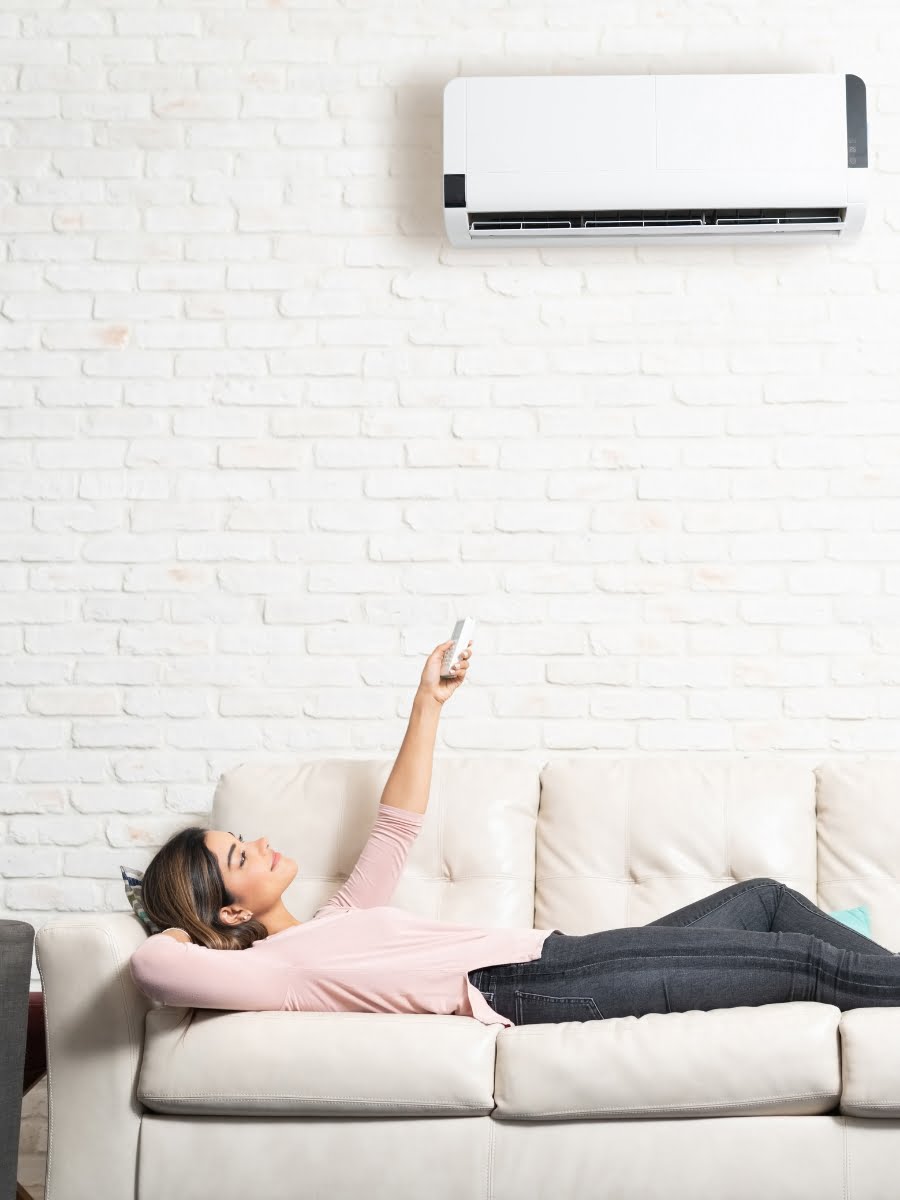 A woman lies on a cream-colored sofa with her head resting on her arm, pointing a remote control towards a ductless mini split air conditioner mounted on a white brick wall above her.