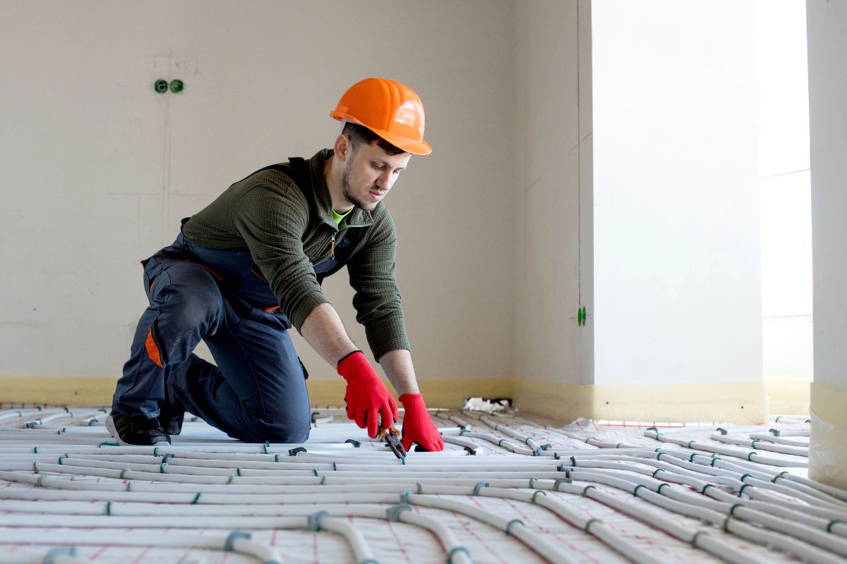 A construction worker wearing an orange hard hat and red gloves installs pipes for hot water recirculation systems on the floor of a room.