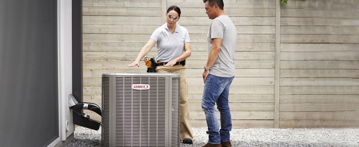 A technician explains a Lennox-branded outdoor air conditioning unit to a man near a house with a grey wall and a wooden fence, while also discussing the benefits of timely furnace repair for optimal home comfort.