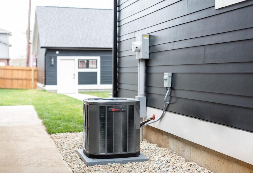 An outdoor air conditioning unit, perfectly installed next to a house with dark siding, sits on a concrete pad surrounded by gravel. A small outbuilding and wooden fence add to the backdrop. Regular air conditioning repair can help maintain its efficiency and keep your cool intact.