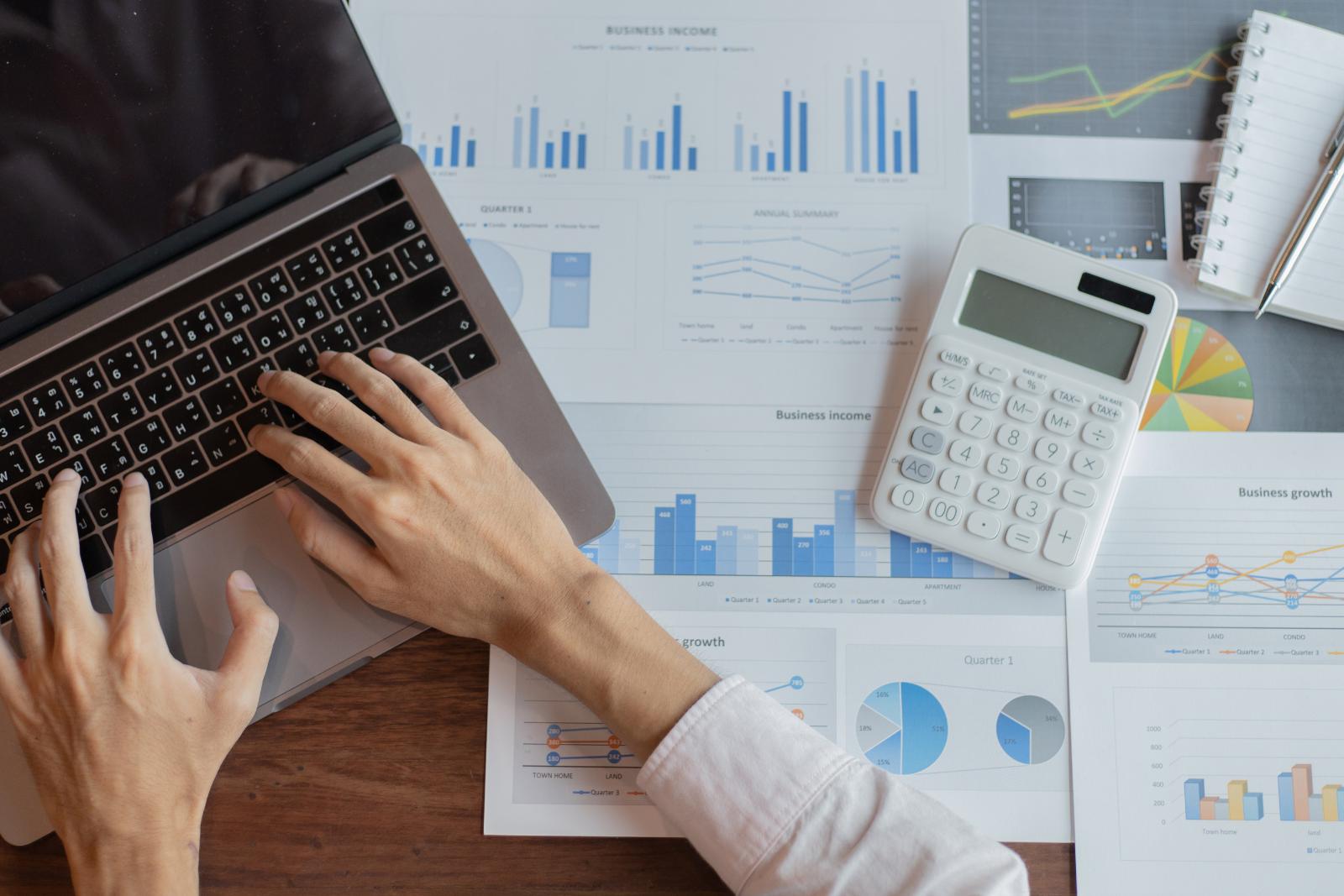 Hands typing on a laptop keyboard next to financial charts, graphs, a calculator, and a notebook on a desk, ensuring the maintenance of accurate records.