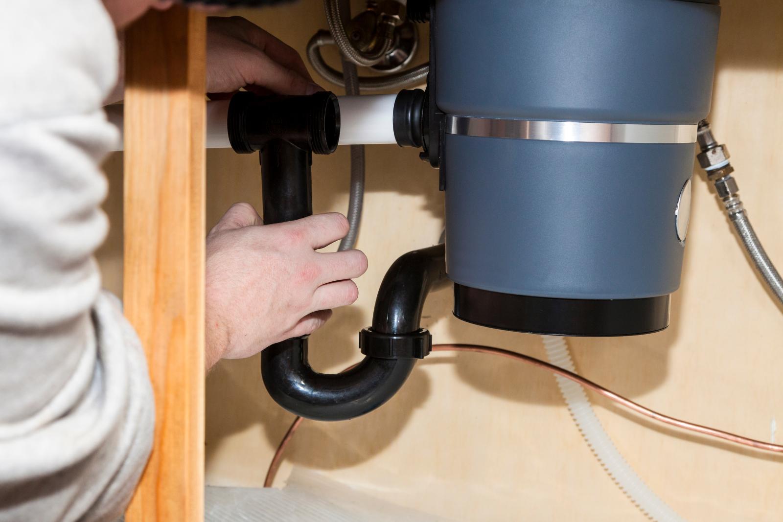 A person is installing or repairing a garbage disposal unit under a kitchen sink, providing expert plumbing services with hands on the black plumbing pipes connected to it.