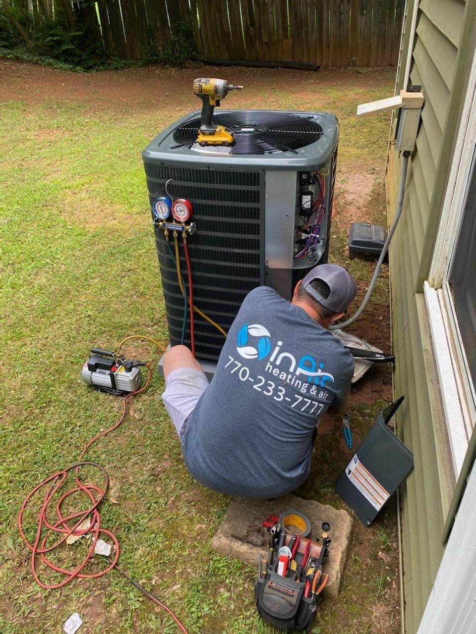 A technician kneels beside an outdoor air conditioning unit, performing maintenance as part of comprehensive HVAC services. Various tools and equipment are placed around him. The technician wears a shirt with a logo and contact number on the back.