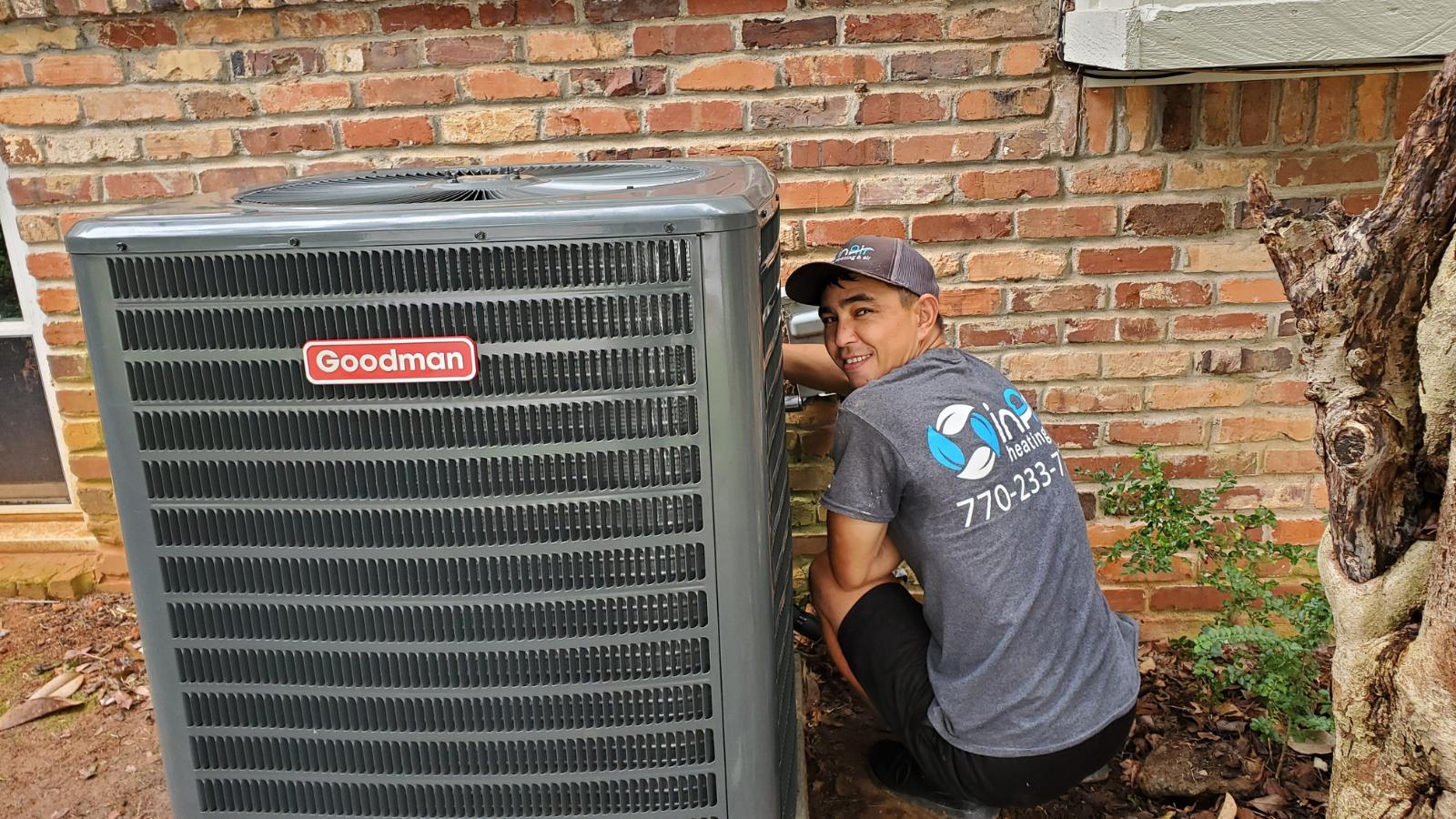 A technician in a gray shirt and cap is working on a Goodman air conditioning unit outside a brick building, ensuring the Service Areas are well-maintained.