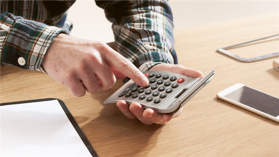 Person in a plaid shirt using a calculator on a wooden desk, focusing intently on financing calculations with a notepad and smartphone nearby.