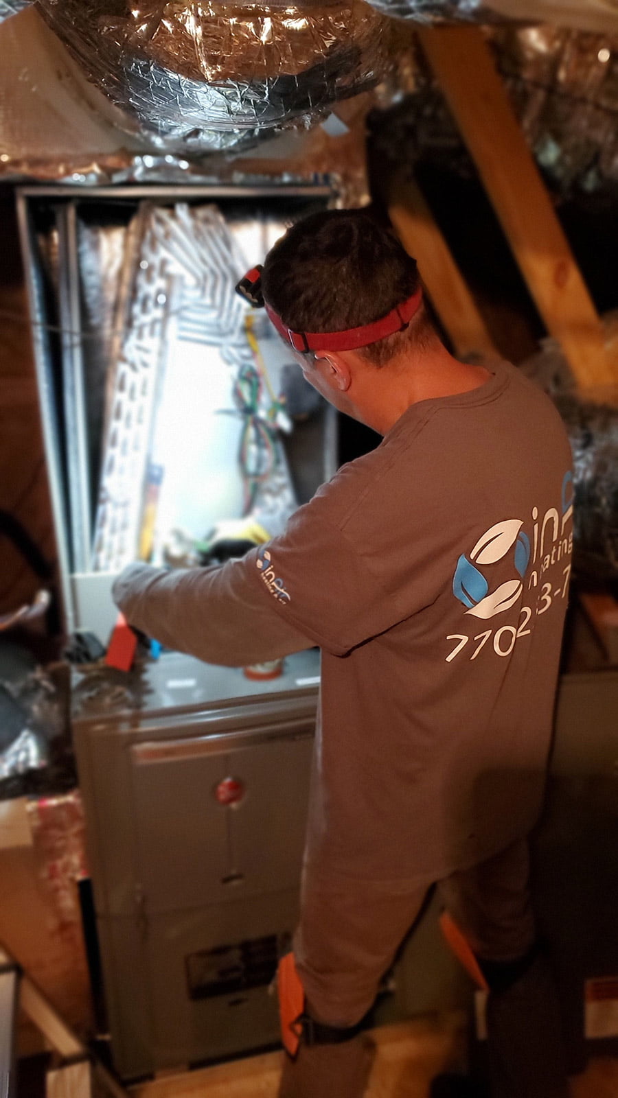 A technician wearing a headlamp and branded shirt works on an open HVAC unit in an attic, surrounded by ductwork and insulation, ensuring top-quality Atlanta HVAC services.