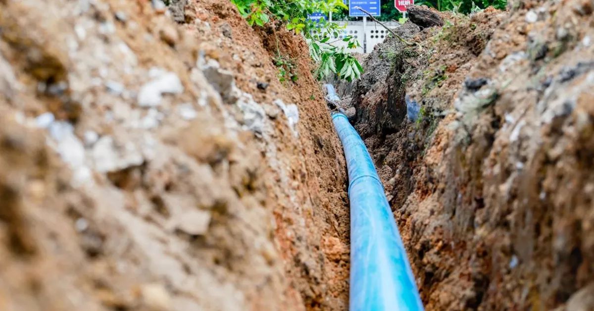 A blue plastic pipe, part of vital plumbing services, is laid in a trench dug through dirt and soil, surrounded by green foliage.