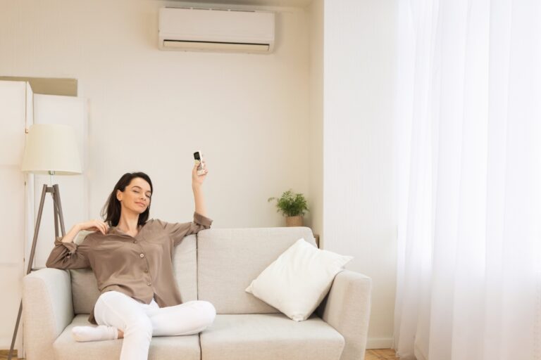 A person is sitting on a couch with a remote control in hand, facing an affordable air conditioner mounted on the wall. The room has light-colored decor and a floor lamp.