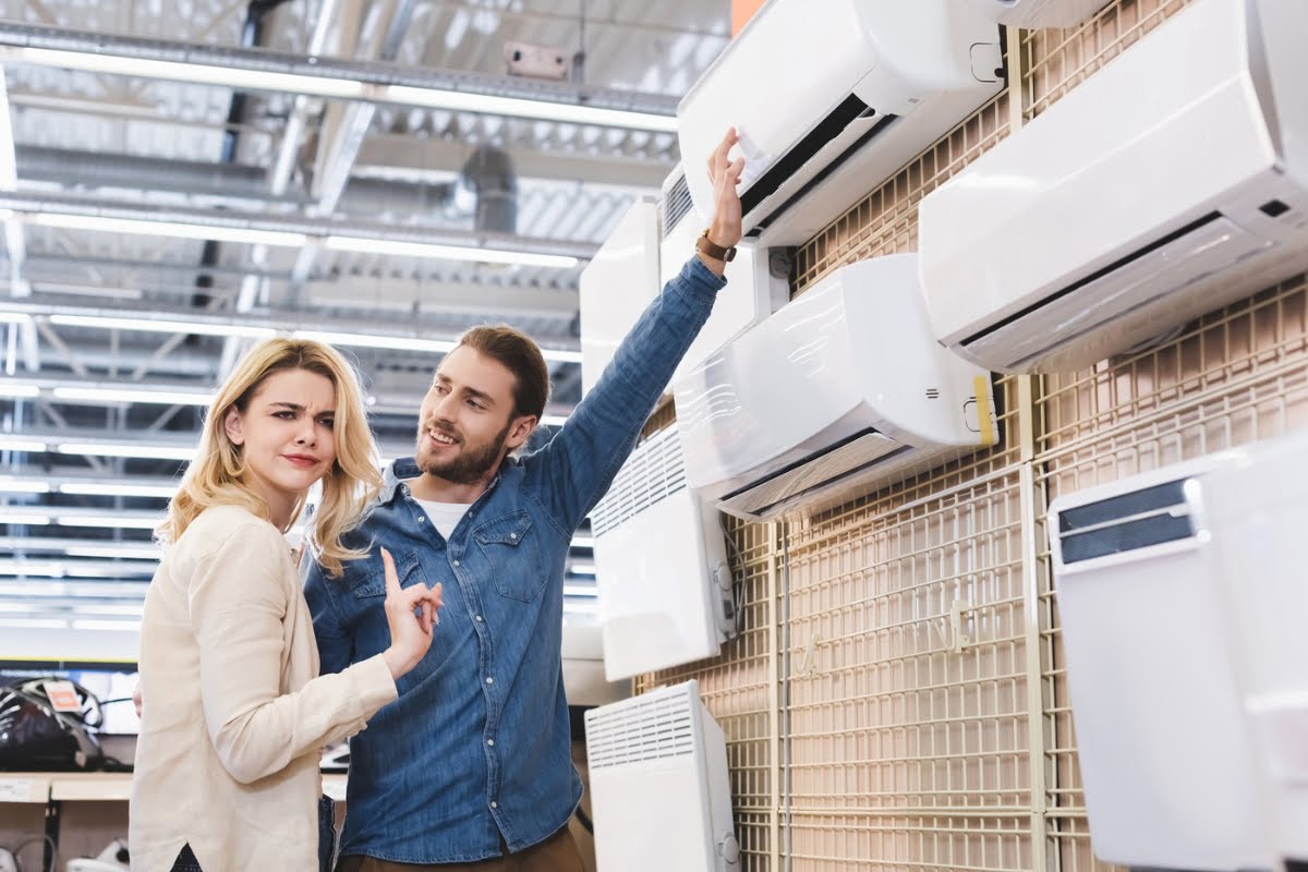 A woman and a man inspect a row of affordable air conditioning units mounted on a display wall in a store. The man is pointing towards one of the units while the woman is looking back with a concerned expression.