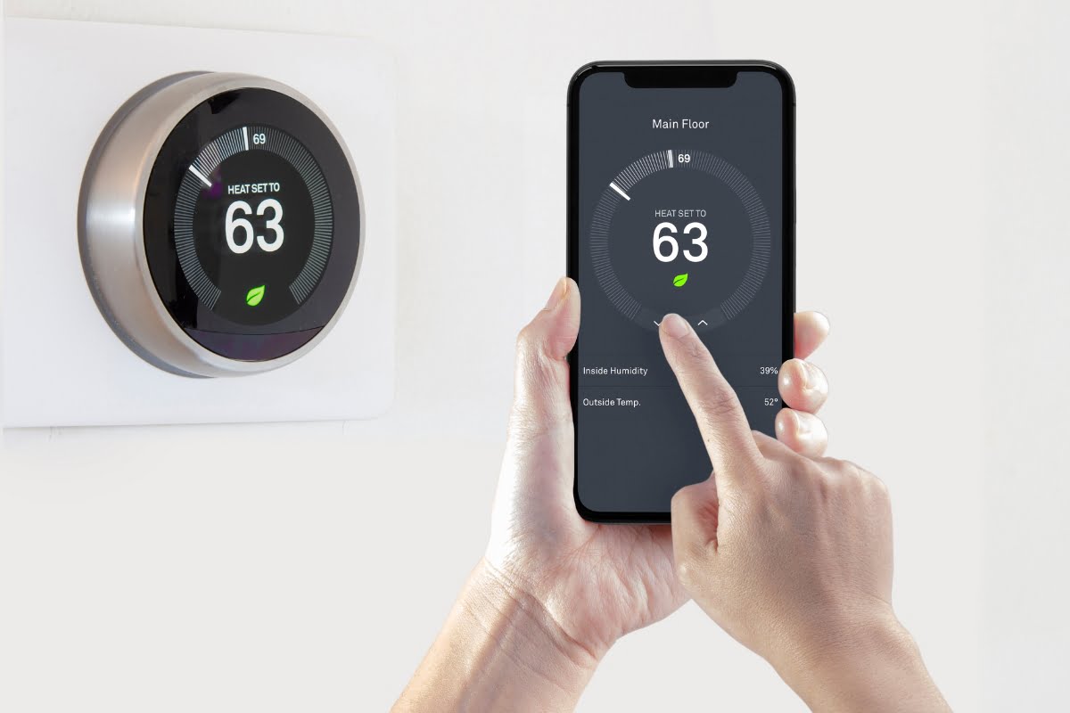 A person adjusts a smart thermostat set to 63°F via a smartphone app. The thermostat and app show current temperature, indoor humidity, and outside temperature. This setup offers an efficient and affordable air conditioning solution for maintaining comfort.