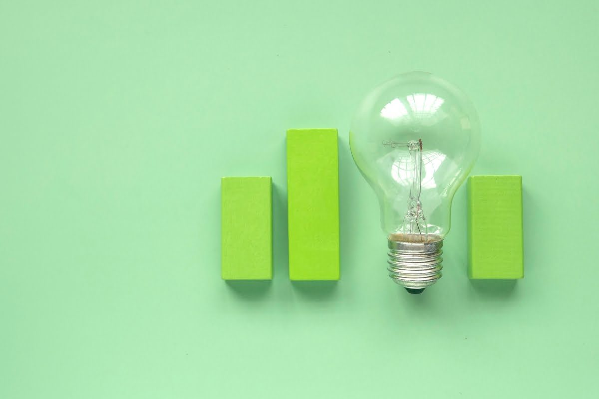 A light bulb is placed next to three green rectangular blocks of different heights on a green background, much like an arrangement showcasing affordable air conditioning units in a sleek, modern design.