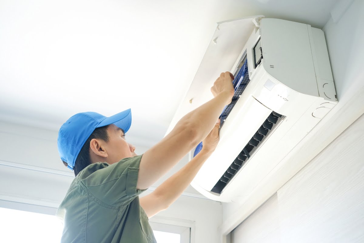 A person wearing a blue cap and green shirt is inspecting or repairing an affordable air conditioning unit mounted on a wall.