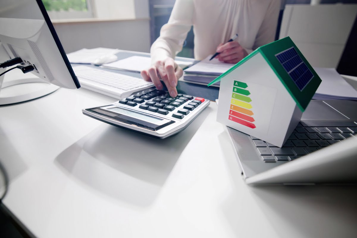 Person using a calculator at a desk with a small model house displaying an energy efficiency chart, and a computer keyboard visible in the background. The setup hints at the importance of affordable air conditioning in maintaining energy-efficient homes.