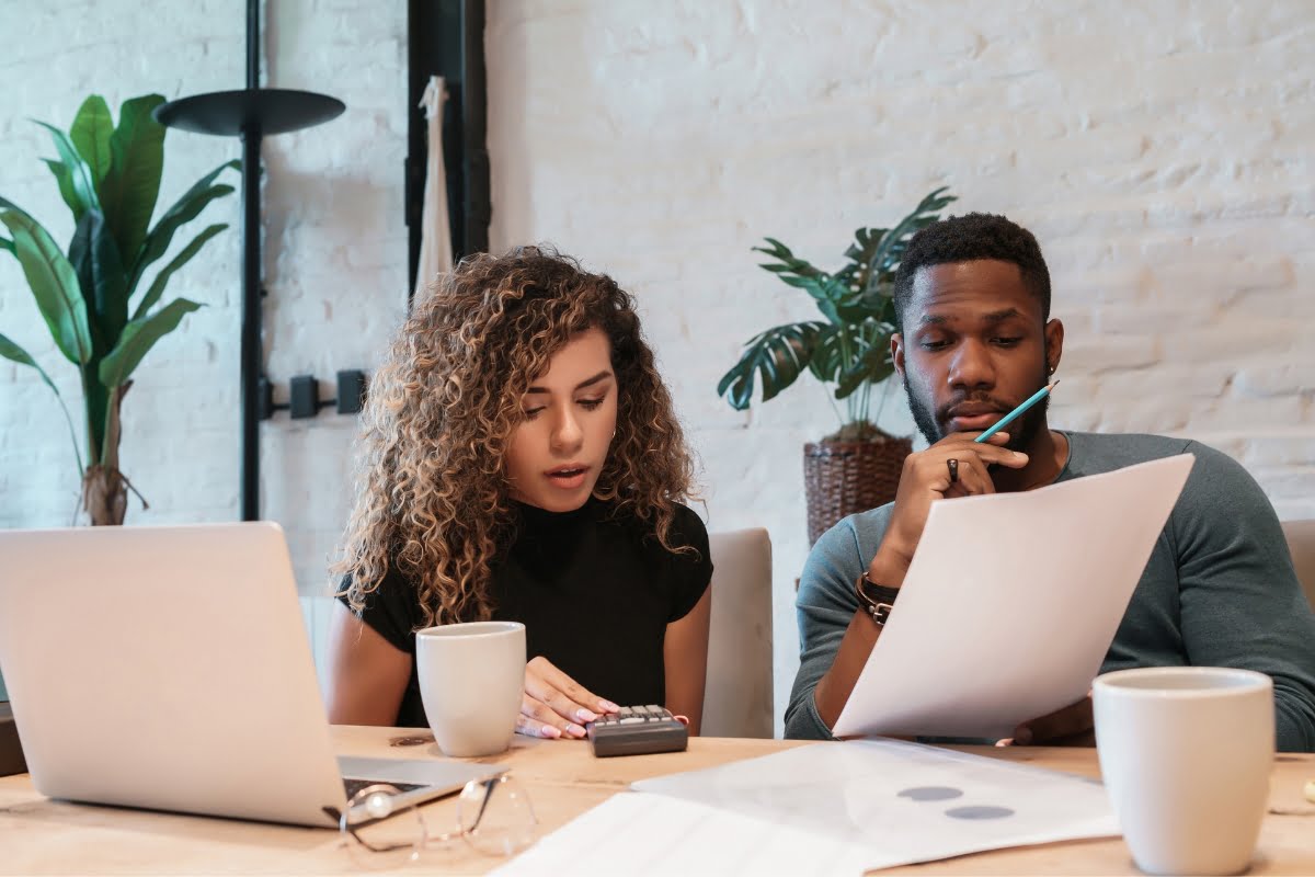 Two people are working at a table with a laptop, papers, a calculator, and coffee cups in front of them. They appear to be focused on their work, perhaps choosing the right HVAC contractor for their project.