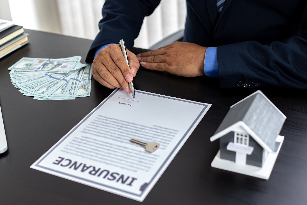 A person is signing an insurance document at a desk with a stack of cash, a key, and a small house model, contemplating the benefits of choosing the right HVAC contractor for their new home.