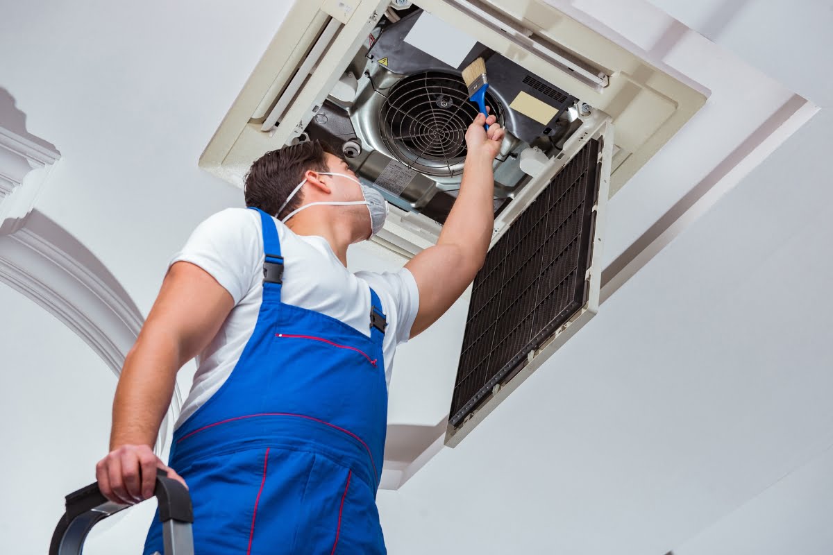 A technician in blue overalls stands on a ladder, performing maintenance on a ceiling-mounted HVAC unit while wearing a face mask, demonstrating the importance of choosing the right HVAC contractor for safety and expertise.