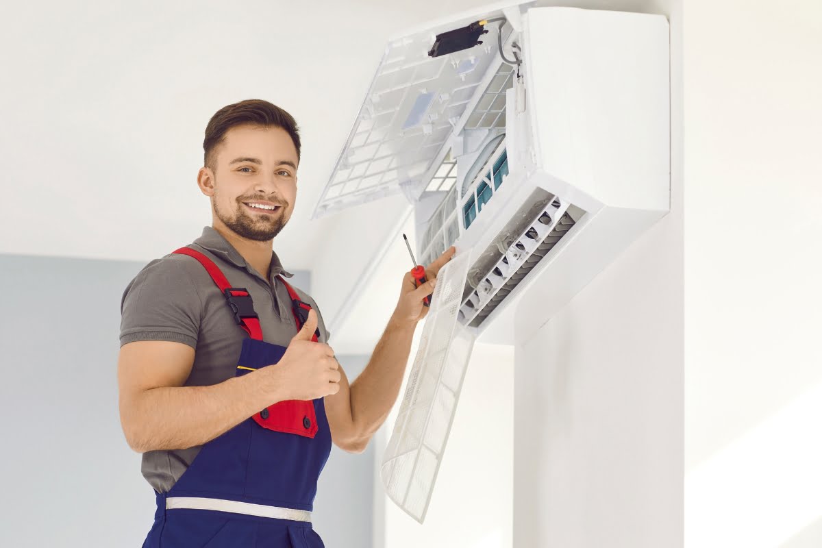 A technician wearing blue overalls and a gray shirt is servicing a wall-mounted air conditioning unit, holding a screwdriver and giving a thumbs-up. Choosing the right HVAC contractor ensures top-notch service like this!