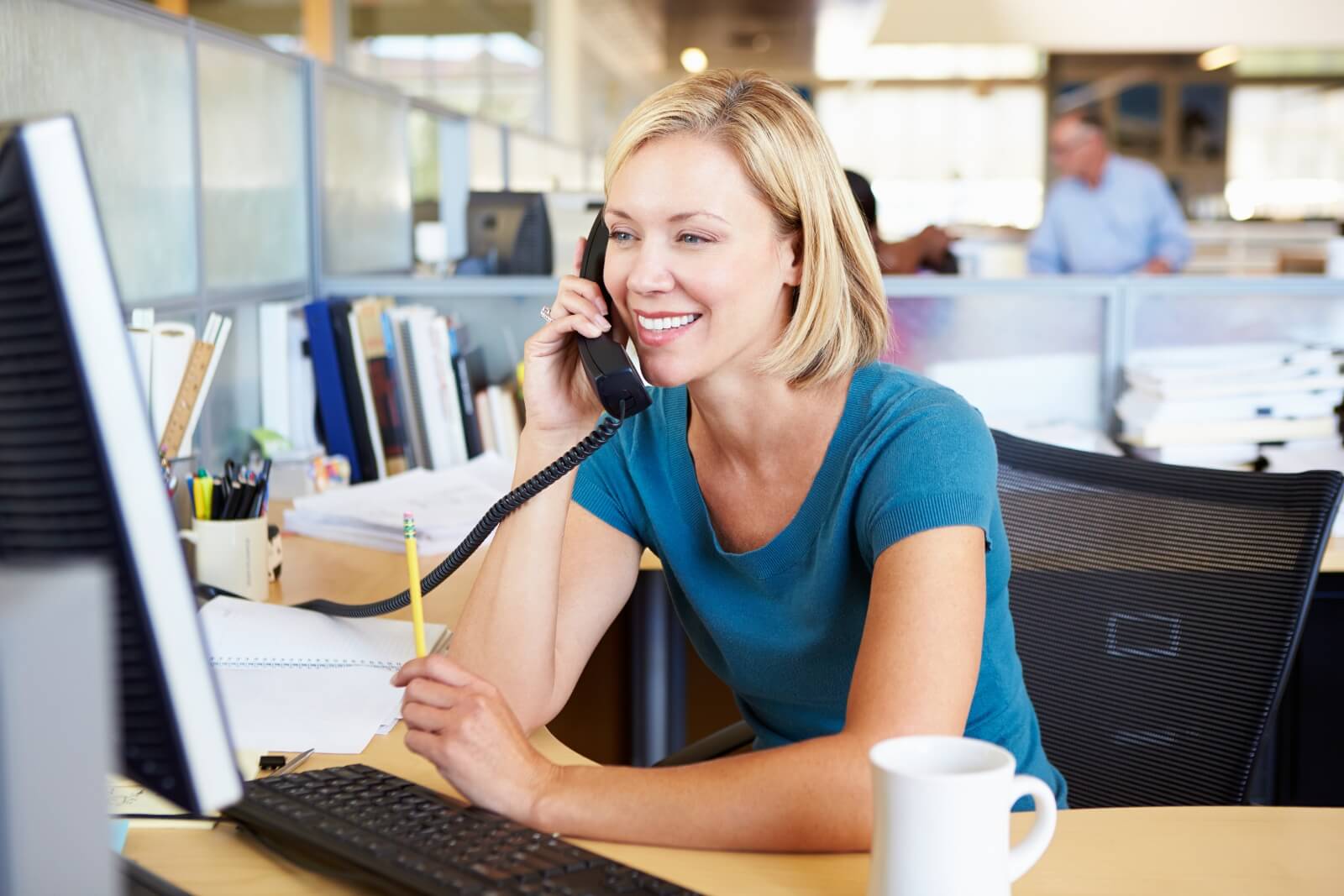 A woman in a blue shirt sits at a desk, talking on the telephone while looking at a computer screen. She holds a pencil as she manages her contact list, and a coffee mug is placed on the desk.