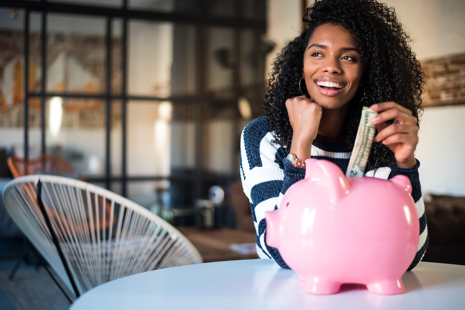 A woman smiles while placing a dollar bill into a pink piggy bank on a table in a modern room with stylish furniture and large windows, embodying the essence of smart financing.