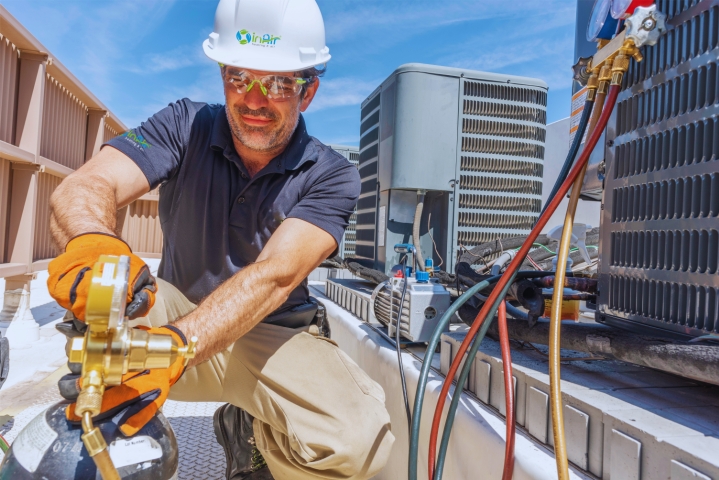 A technician in safety gear installs or repairs an HVAC unit on a rooftop, providing essential HVAC services. He is handling tools and equipment, including a tank and various hoses.