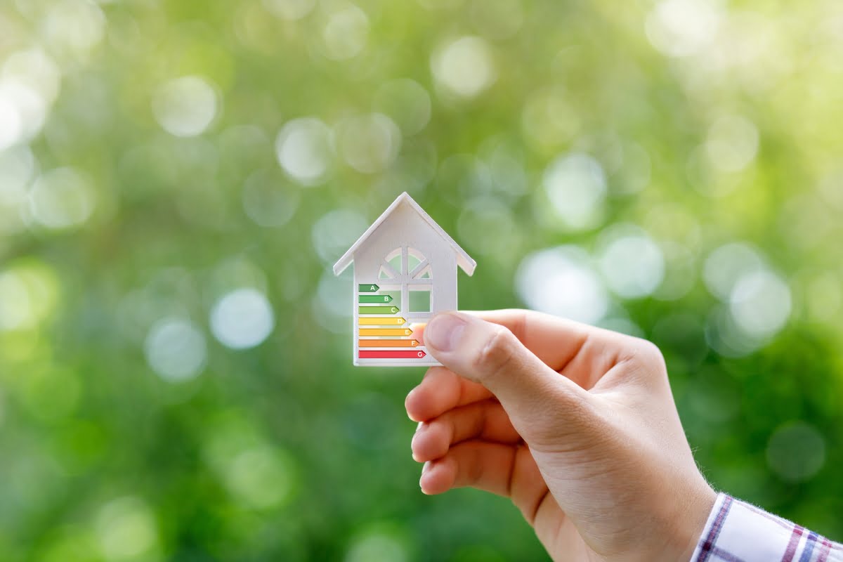 A hand holds a small white house model with a multi-colored energy efficiency rating, reflecting an energy efficient HVAC system. The background is blurred greenery.