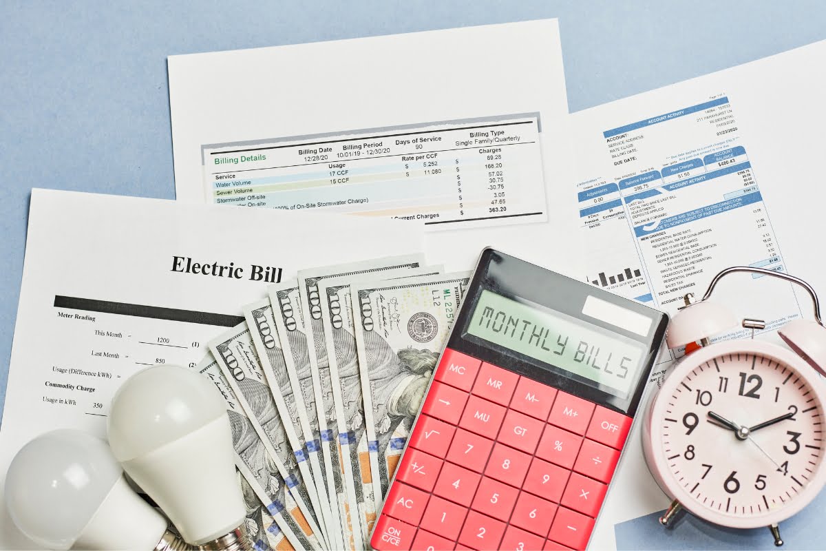 Image showing an electric bill, US dollars, light bulbs, a red calculator with "Monthly Bills" displayed, and a pink alarm clock on a blue background. An energy efficient HVAC system can help reduce the costs shown on the bill.