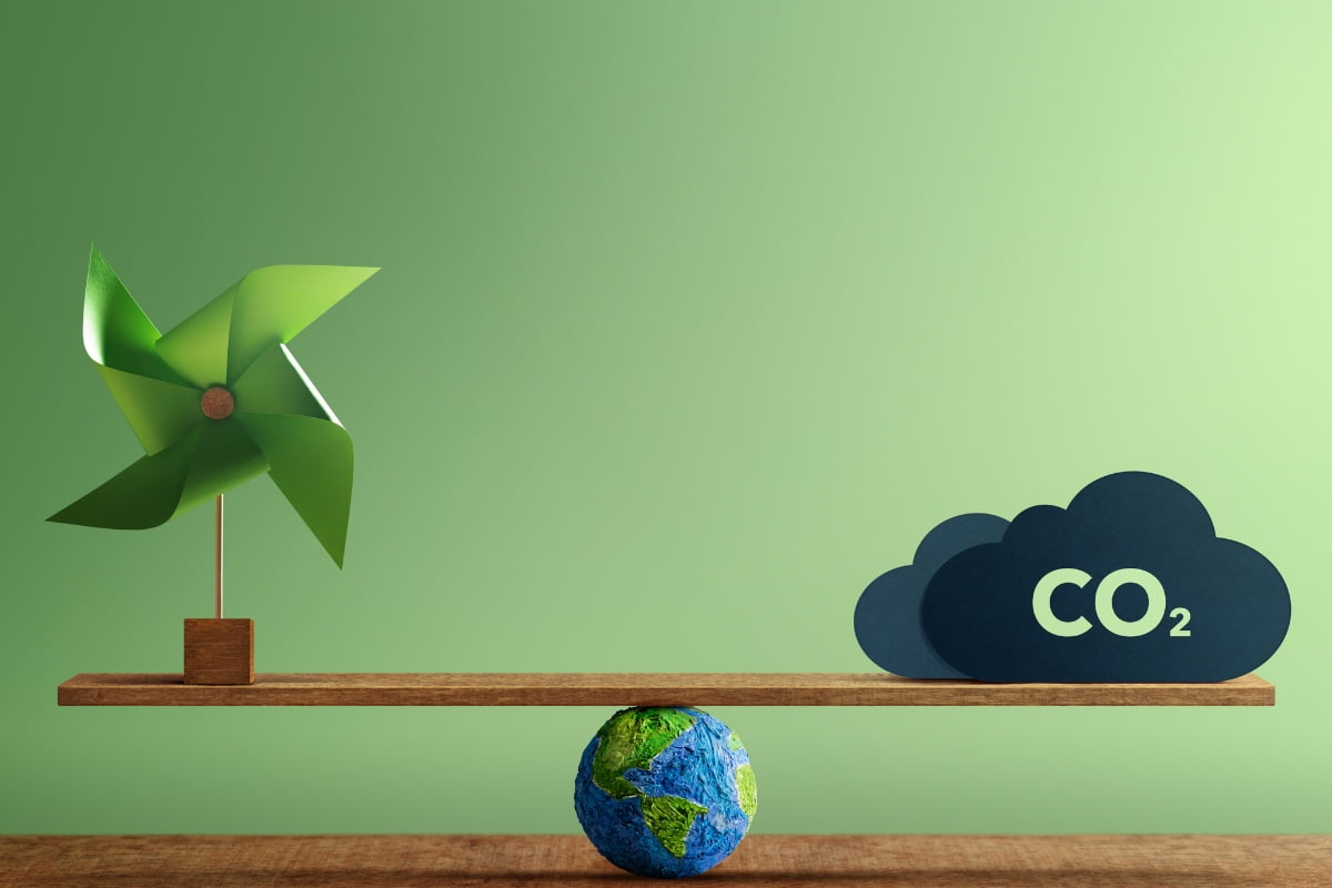 A balance scale with a green pinwheel on the left and CO2 cloud-shaped icons on the right, symbolizing energy efficient HVAC, with a small globe underneath the center. Green background.